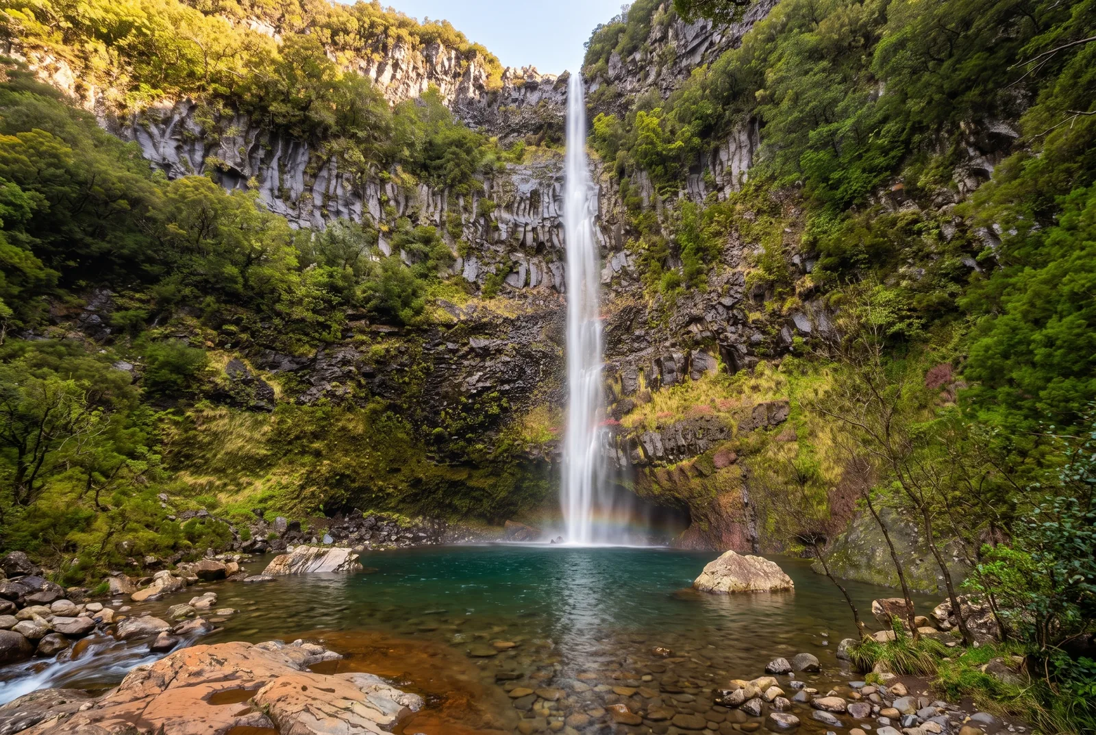 A tall thin waterfall falling into an emerald pool in a green amphitheatre