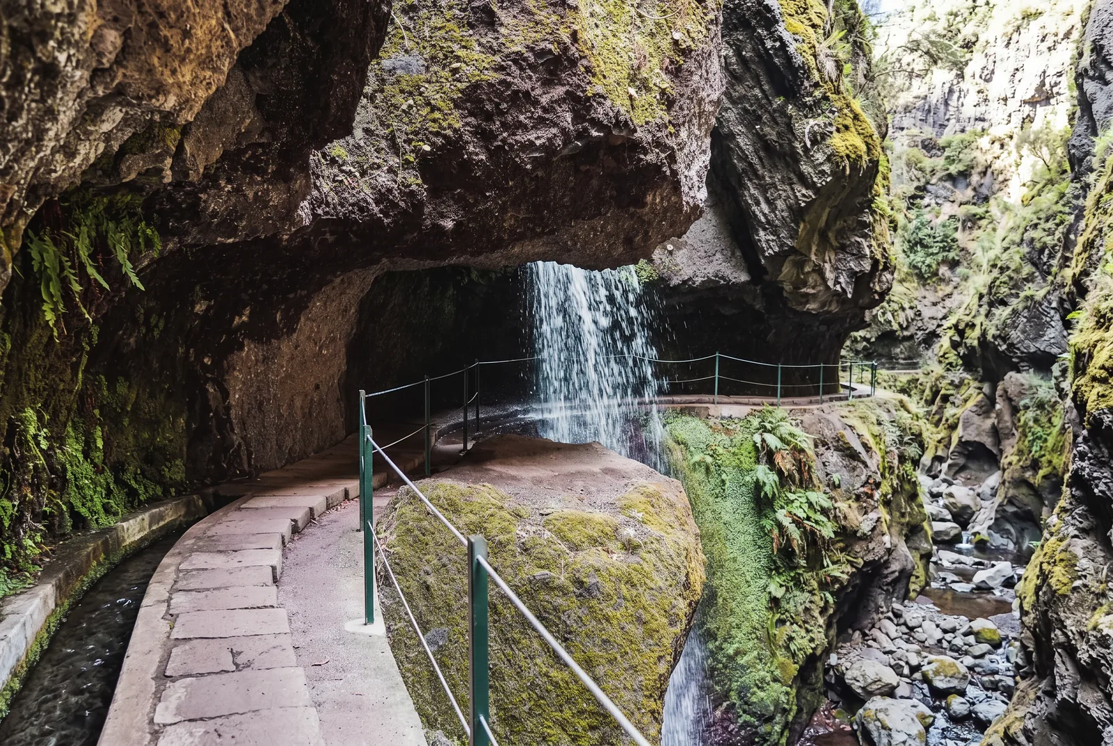 A waterfall pouring over an overhanging rock onto a levada path with a cable railing