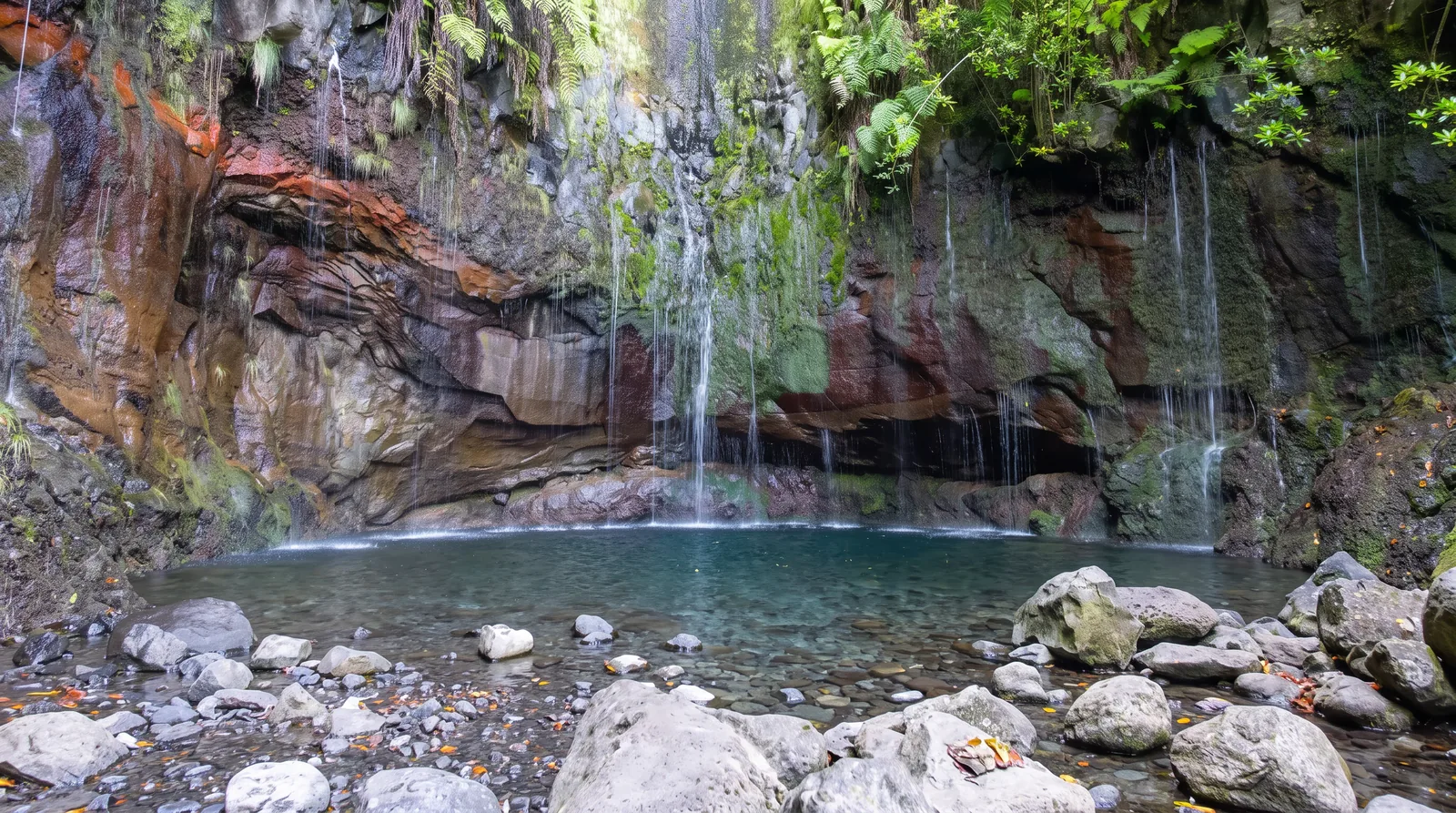 A curved rock amphitheatre with many thin waterfalls dropping into an emerald pool