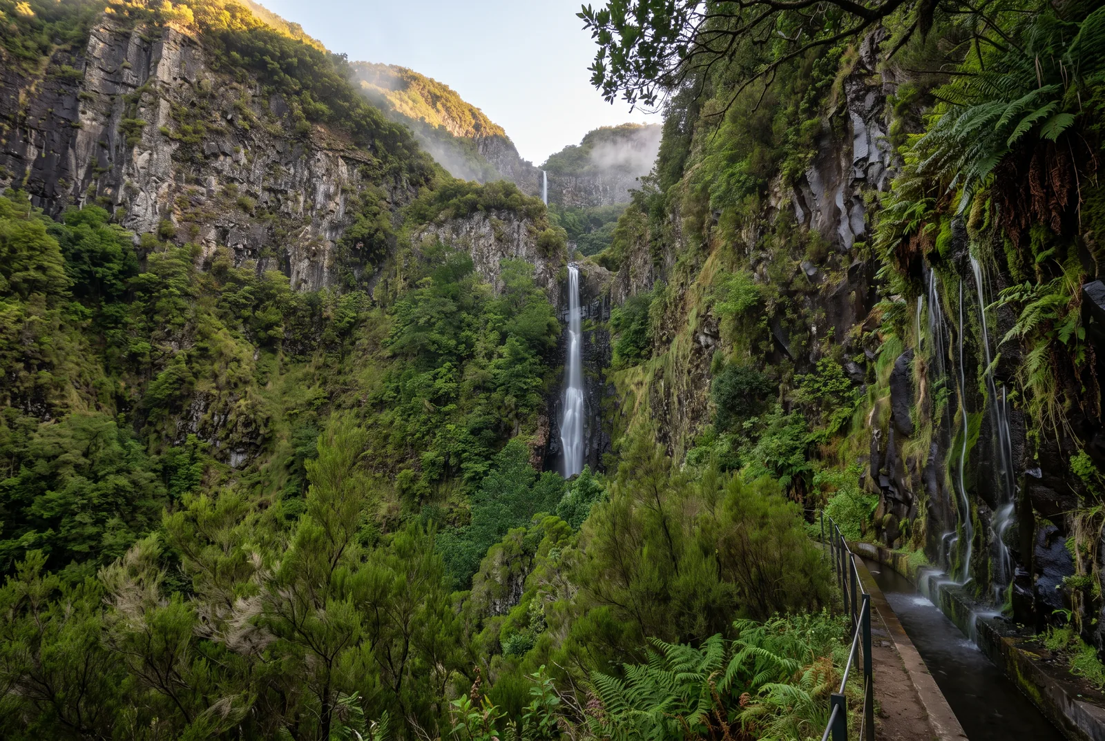 A tall thin waterfall in a green gorge, with a levada channel and railing in the foreground