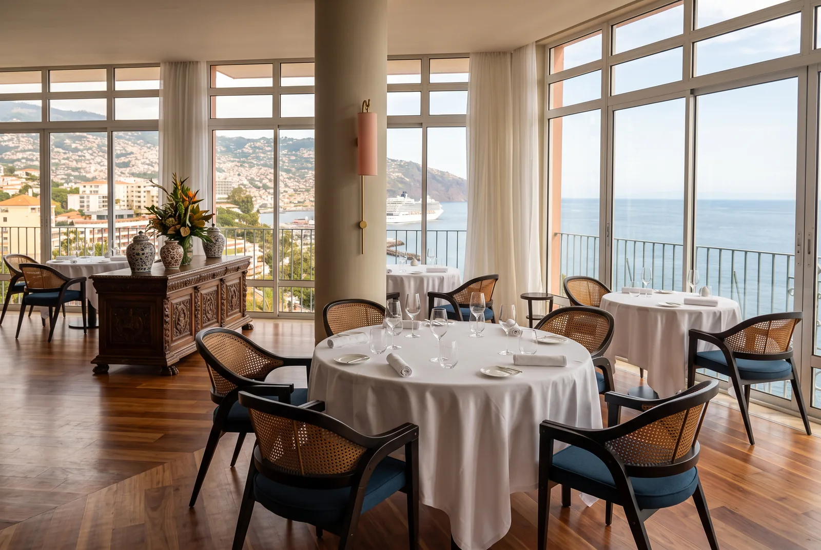 A hotel dining room with laid round tables and floor-to-ceiling windows over Funchal bay