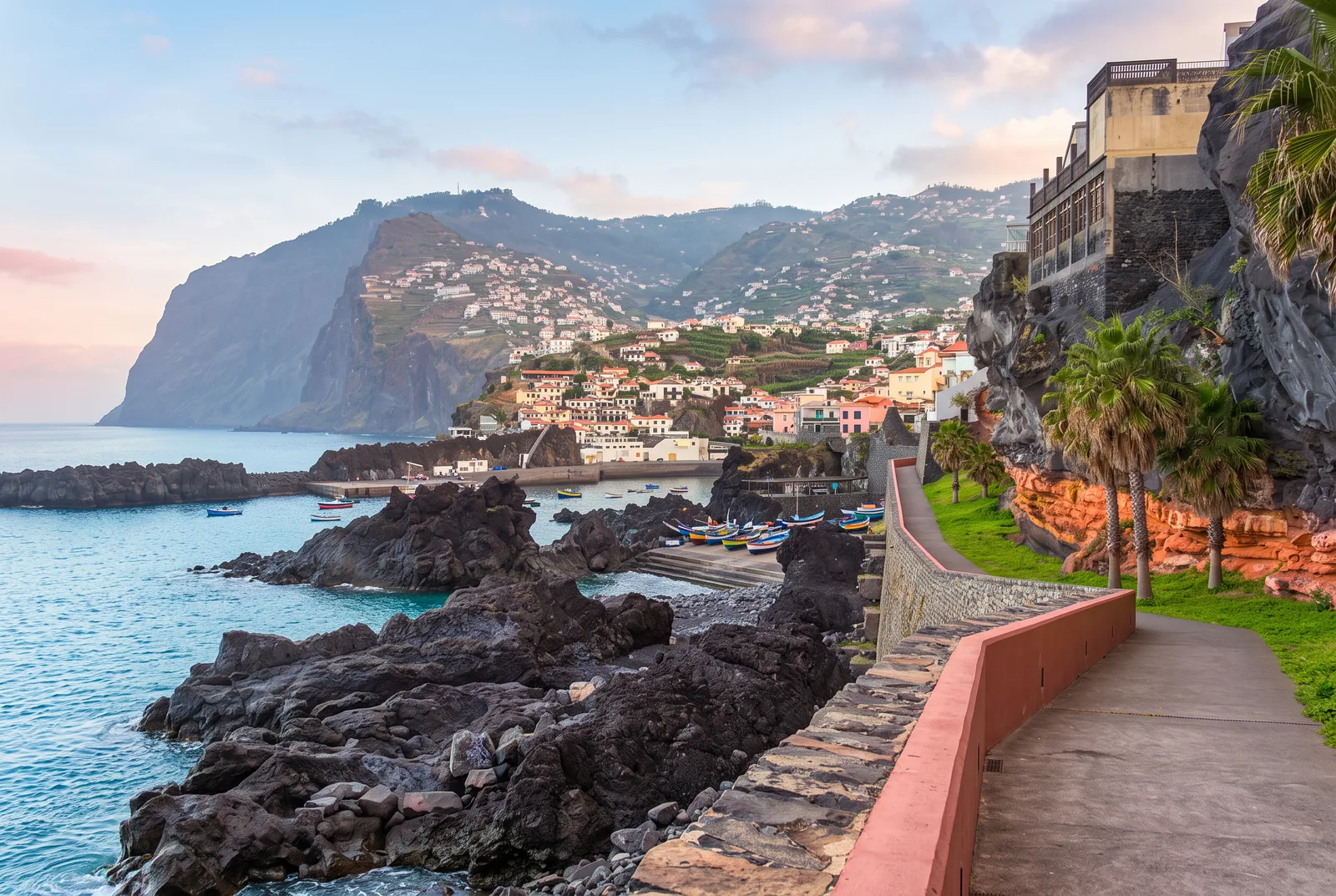 The seaside promenade arriving at Câmara de Lobos, with painted fishing boats and Cabo Girão behind
