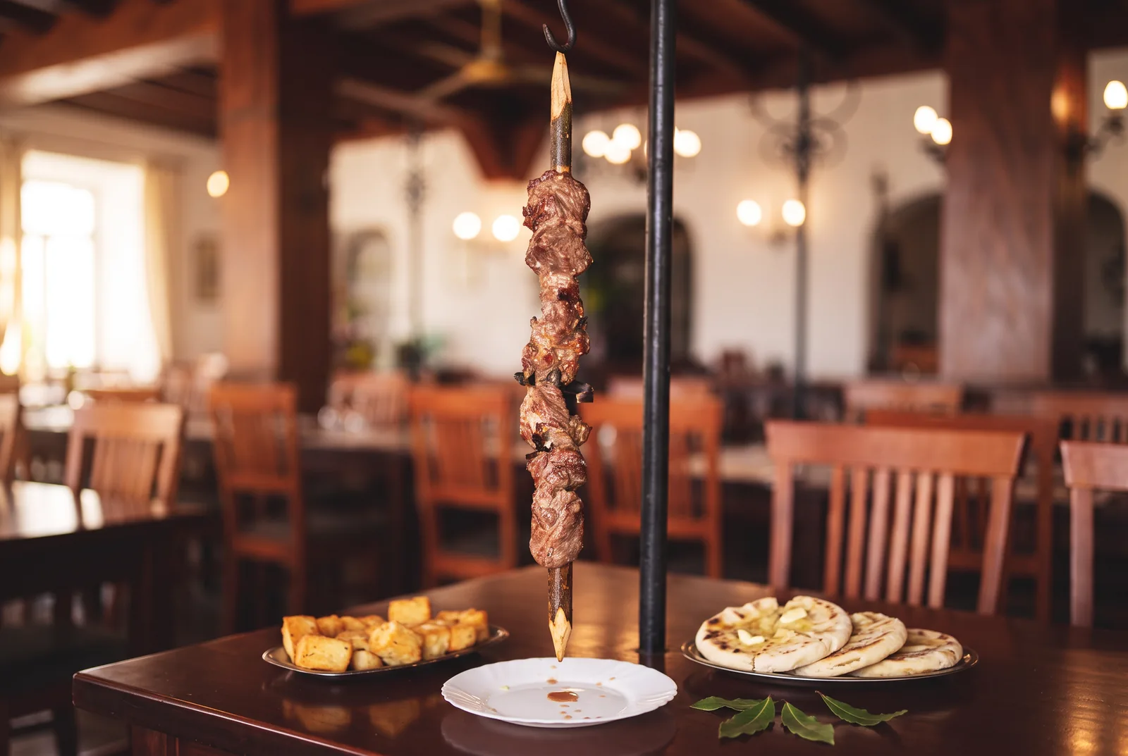 A Madeiran espetada hanging vertically on its stand, beef chunks on a laurel stick with milho frito and bolo do caco alongside