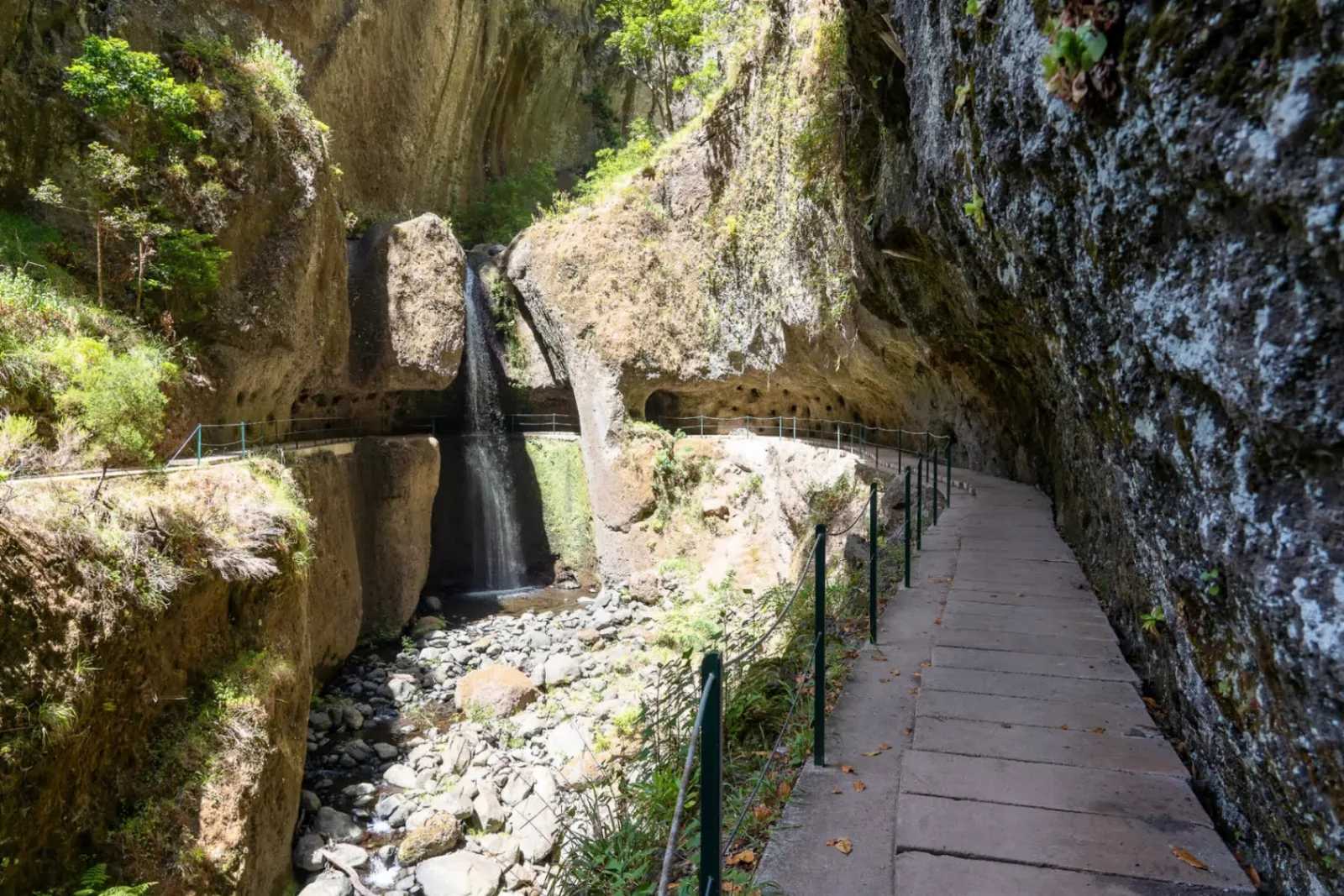 A levada channel running full beside a cliff path, with a waterfall spilling over the rock