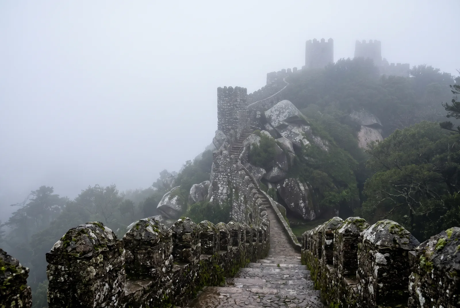 The ramparts of the Moorish Castle in Sintra disappearing into dense mountain fog, with mossy merlons in the foreground