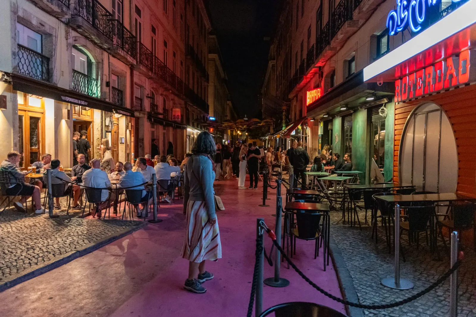 A Lisbon street at night with bar terraces and neon light spilling onto the pavement