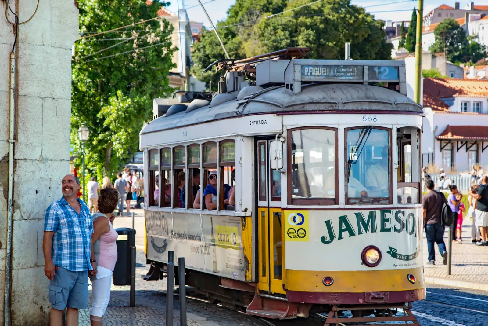 A yellow Lisbon tram waiting at a stop on a leafy street