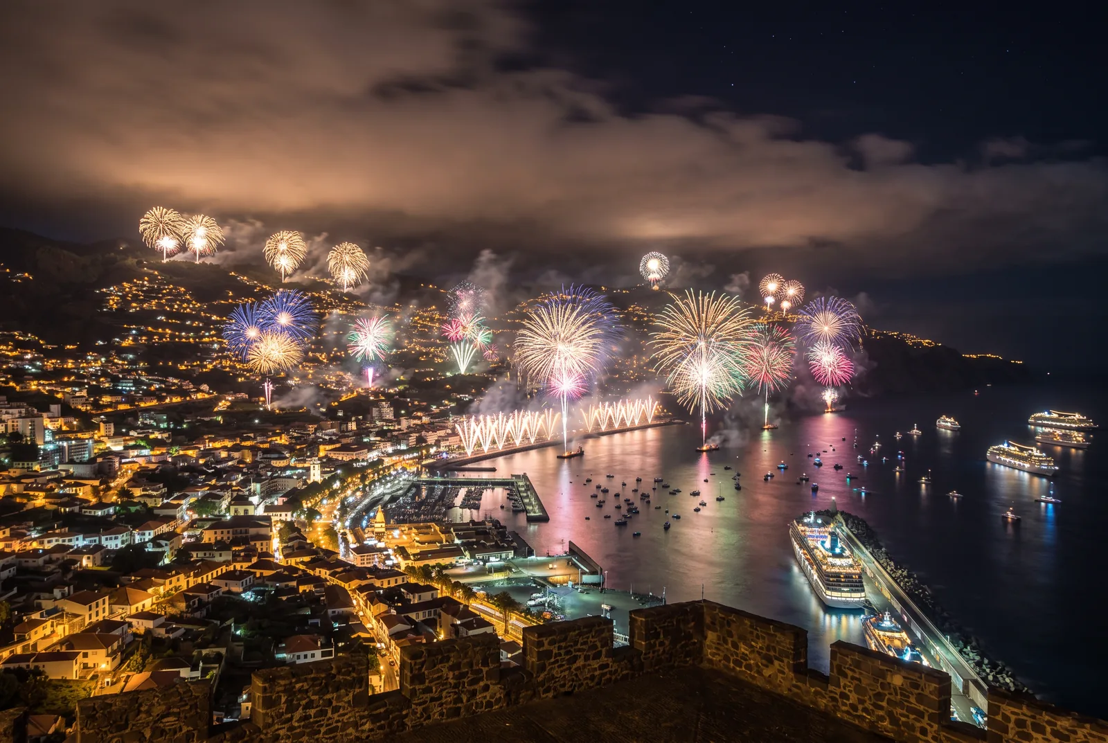 Fireworks bursting over the Funchal amphitheatre and harbour on New Year's Eve, with illuminated cruise ships moored at the Pontinha breakwater and anchored in the bay