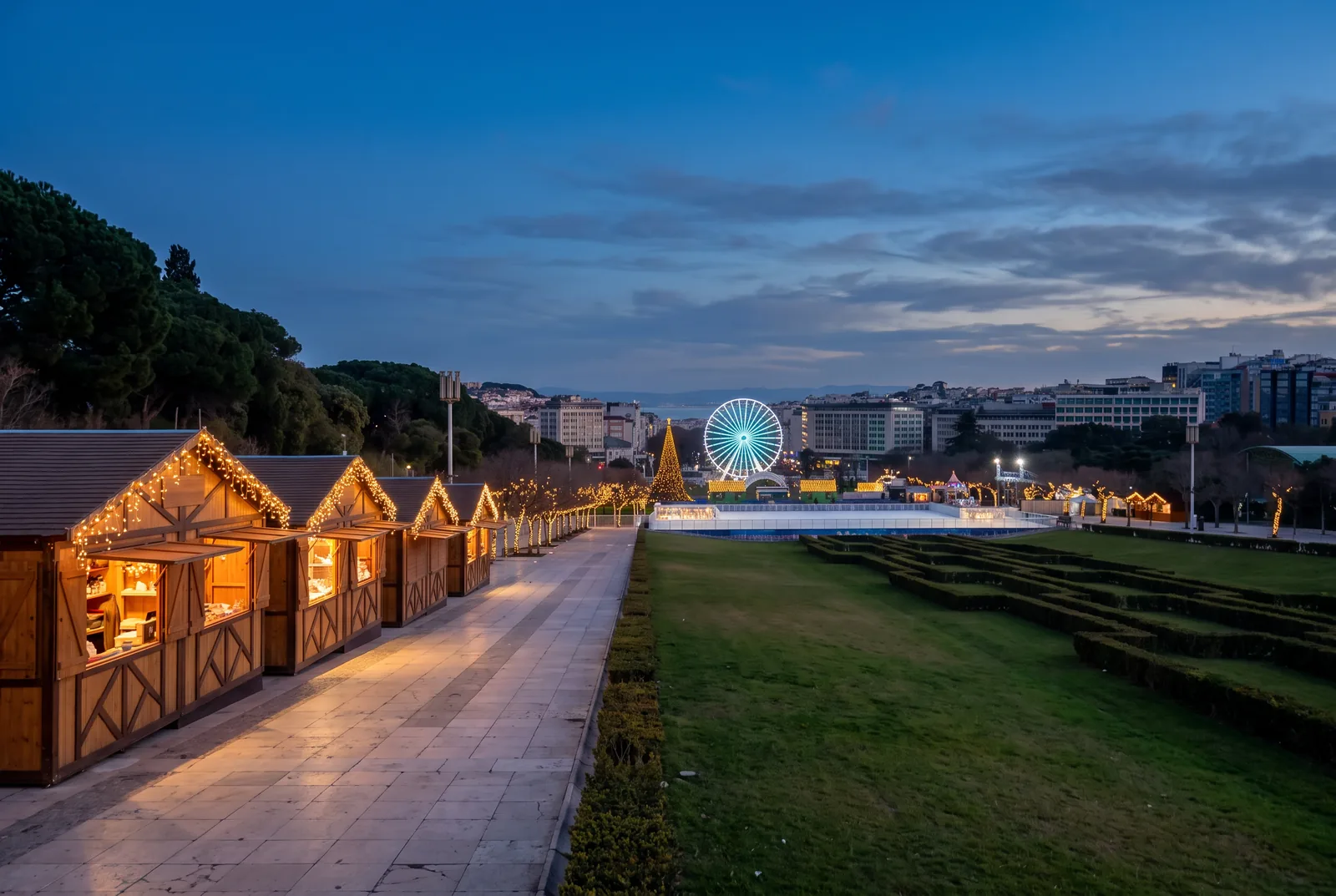 Wooden Christmas stalls lit along a promenade in Parque Eduardo VII, with the Ferris wheel and the Tagus beyond