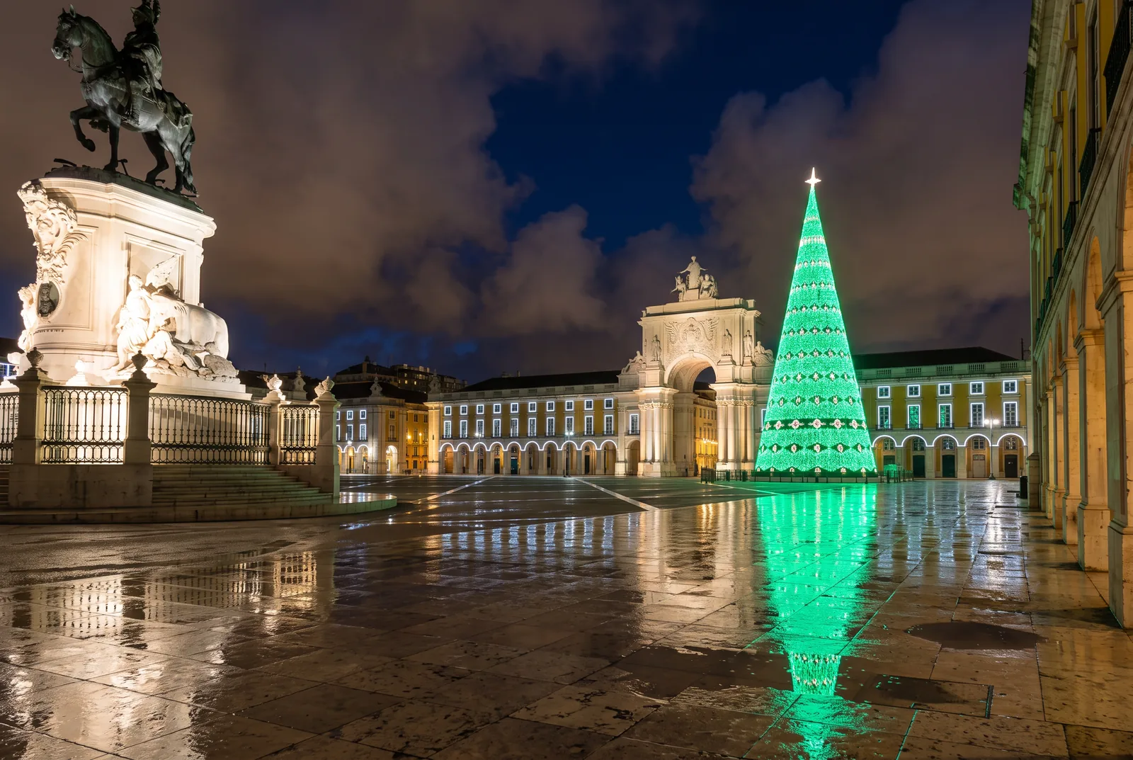 The giant illuminated Christmas tree in Praça do Comércio at night after rain, with the Arco da Rua Augusta and the statue of King José I