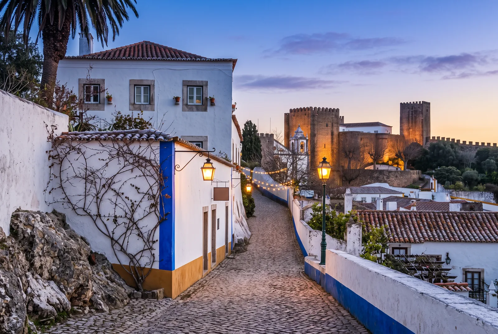 A cobbled lane inside the walls of Óbidos at dusk with Christmas lights strung across it and the castle lit above the rooftops