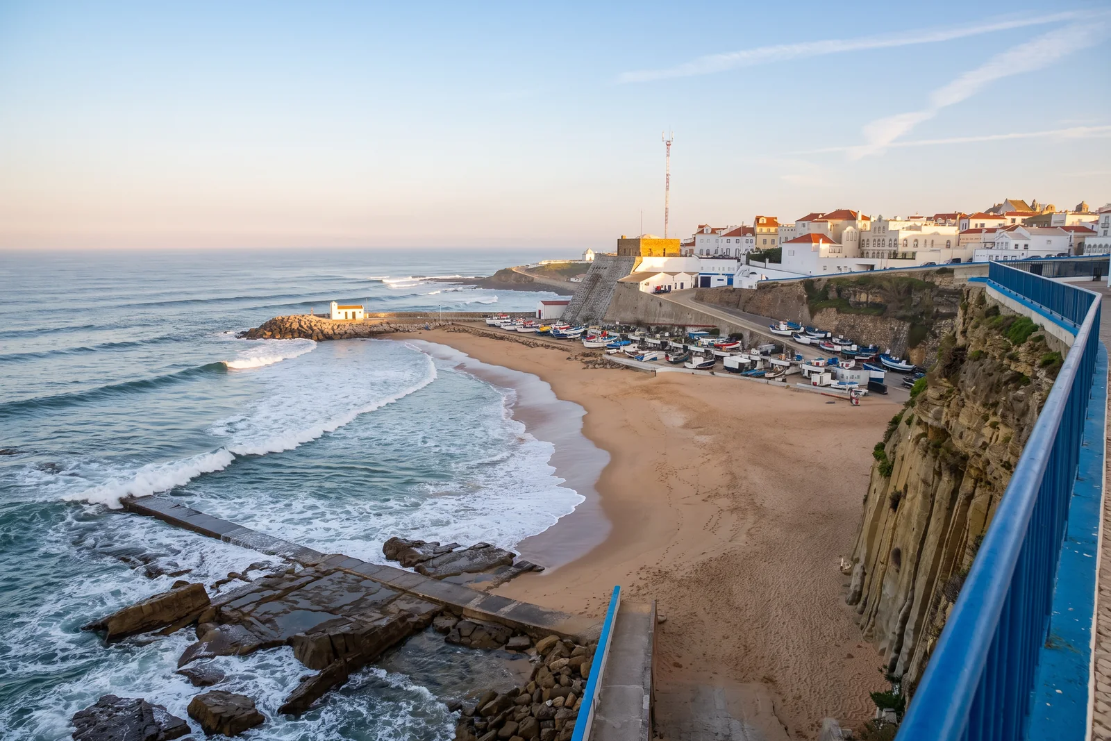 Praia dos Pescadores in Ericeira seen from the clifftop promenade, with fishing boats on the harbour ramp and whitewashed houses along the cliff