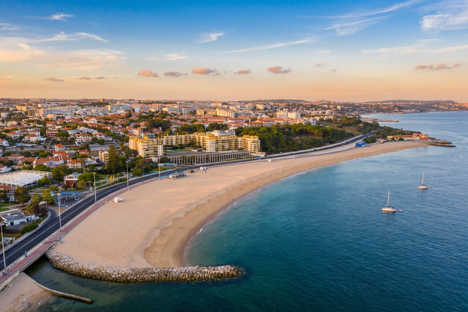 Praia de Santo Amaro de Oeiras from above at golden hour, the crescent of sand curving from the rock groyne past the seafront promenade, with sailing boats anchored in the Tagus estuary