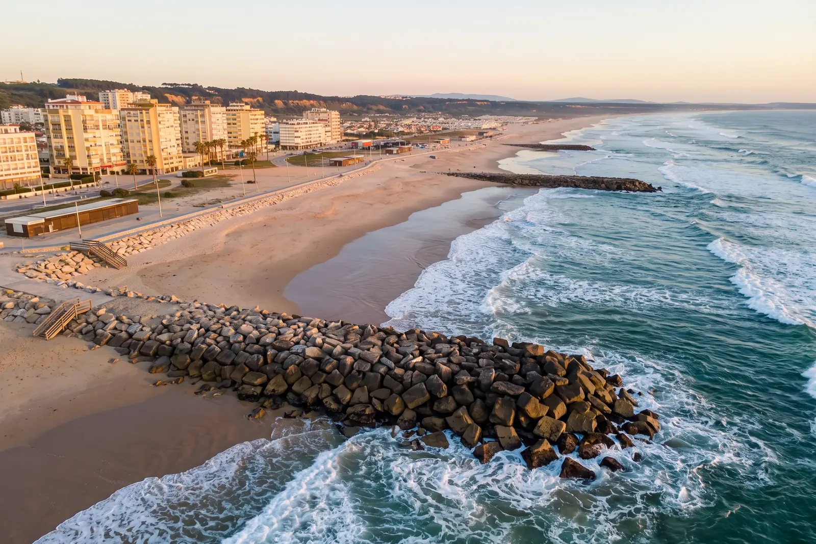 The urban beachfront of Costa da Caparica at golden hour, rock groynes reaching into the Atlantic surf, a wide empty beach and apartment blocks along the promenade