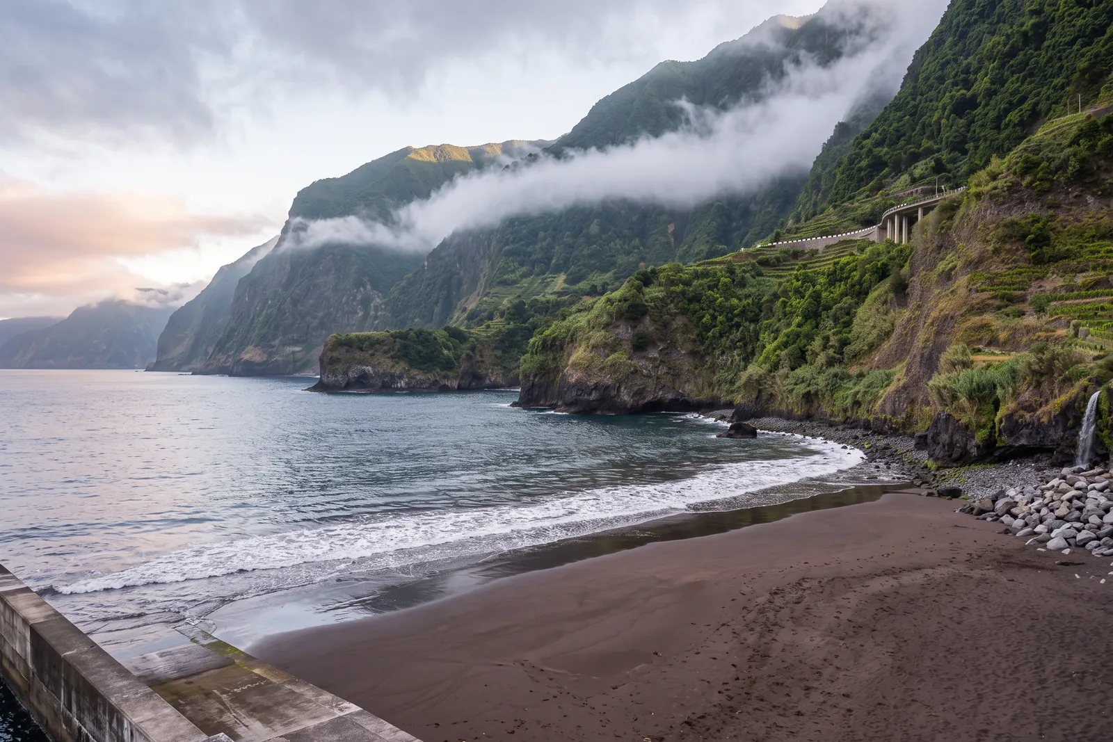 The black volcanic sand beach at Seixal on Madeira's north coast at dawn, with steep green cliffs, the coastal road viaduct and low cloud on the slopes