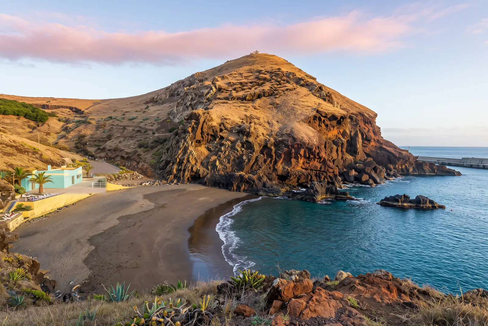 Prainha do Caniçal in early morning, a small dark volcanic sand cove below a rust-red basalt headland topped by a white chapel