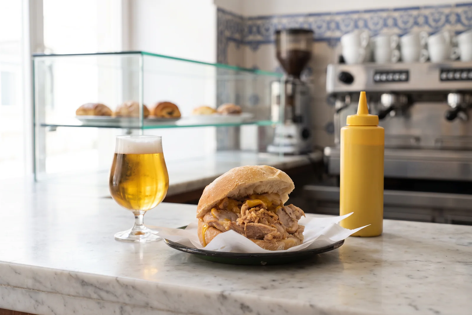 A Lisbon bifana of thin pork slices stewed in white wine and garlic inside a crusty papo seco roll, on greaseproof paper on a marble café counter beside a glass of draught beer and a yellow mustard bottle