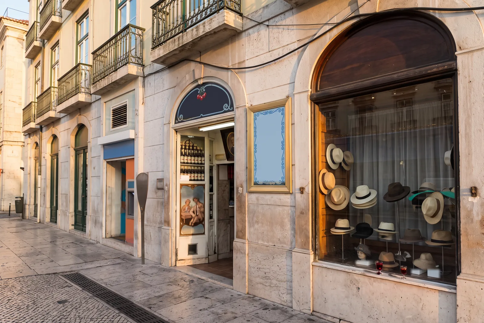 Late-afternoon sun on the arched doorway of A Ginjinha in Largo de São Domingos, Lisbon, with two small glasses of ginjinha resting on the sill of the neighbouring hat shop window